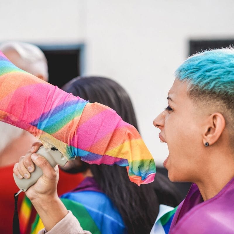A lesbian demonstrator with tan skin and short blue hair, wearing a pride flag wrapped over her shoulders, yelling at a protest into a megaphone wrapped with rainbow fabric.