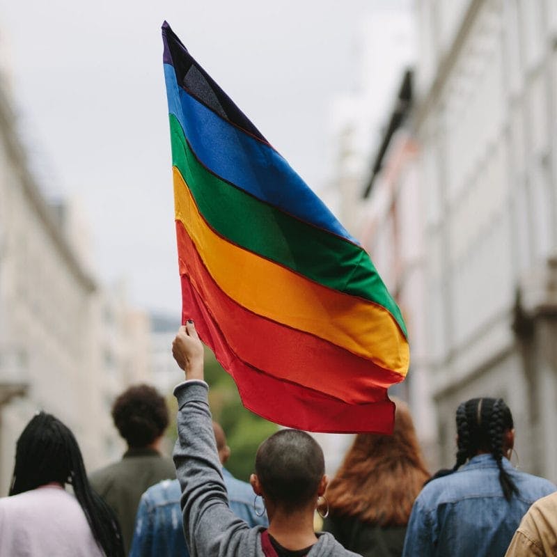 Rear view of young people marching down a city street; the person in foreground holds up a rainbow LGBTQ pride flag
