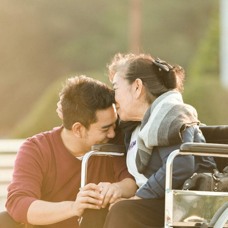 An senior Asian woman is seated in a wheelchair, smiling and embracing a smiling young man sitting beside her chair