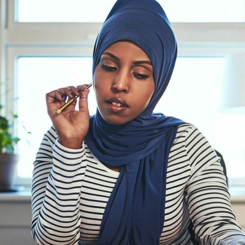 Focused young woman with brown skin wearing a hijab sitting at a desk in her office reading through documents