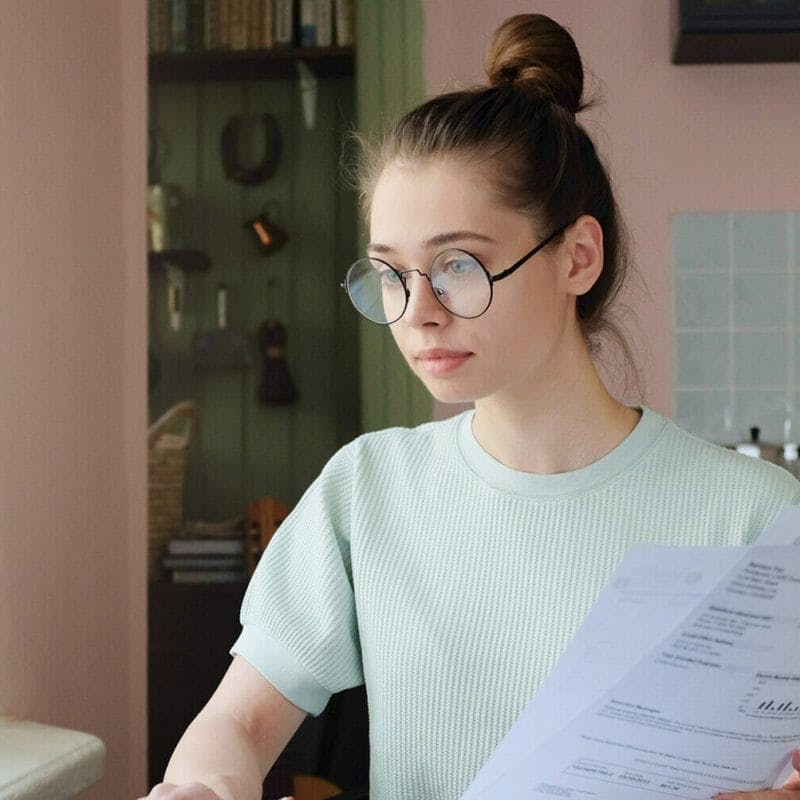 Young woman with fair skin and straight hair in a bun, wearing glasses, sitting at kitchen table with open laptop, bills and calculator, using touchpad, making notes with pencil