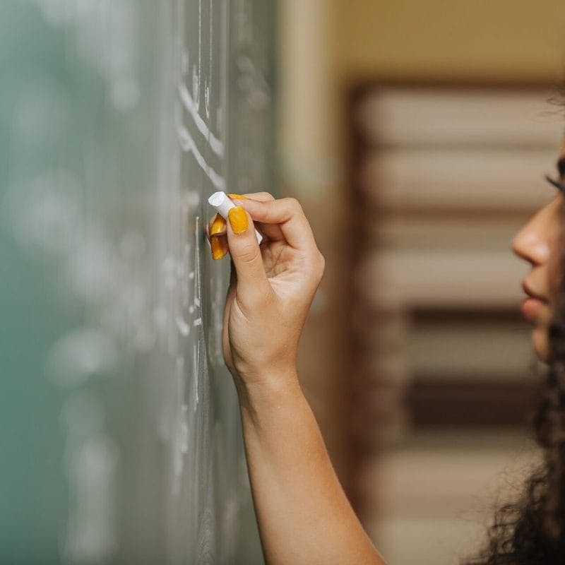 A student with long, curly dark hair and tan skin, wearing bright orange nail polish, writes on a chalkboard.