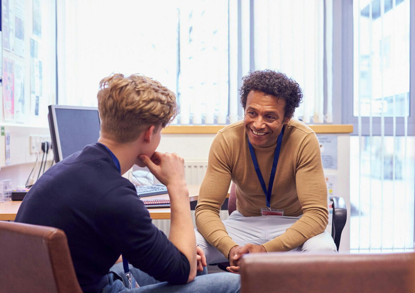 A young person with light skin and short blond hair sits in an office, in conversation with a smiling counselor with brown skin and curly brown hair.