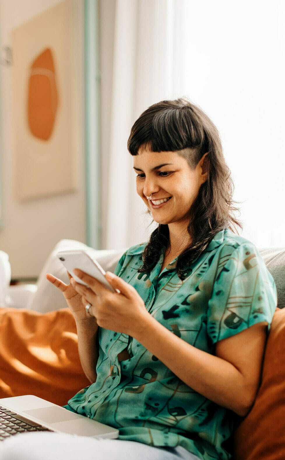 Young woman reading a text message on her smartphone. Happy young woman smiling cheerfully while holding a smartphone on her couch. Young woman sitting with a laptop in her living room.